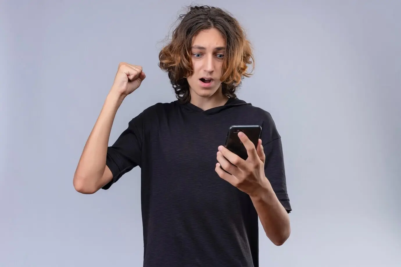 Удивлённый длинноволосый парень в чёрной футболке держит телефон на белом фоне.